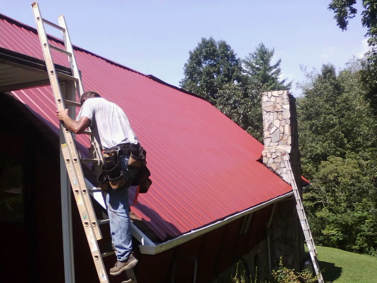 Roofer on a ladder beside a red metal roof and stone chimney — Soffit & Fascia Repair by Roofing Brothers in Stanley